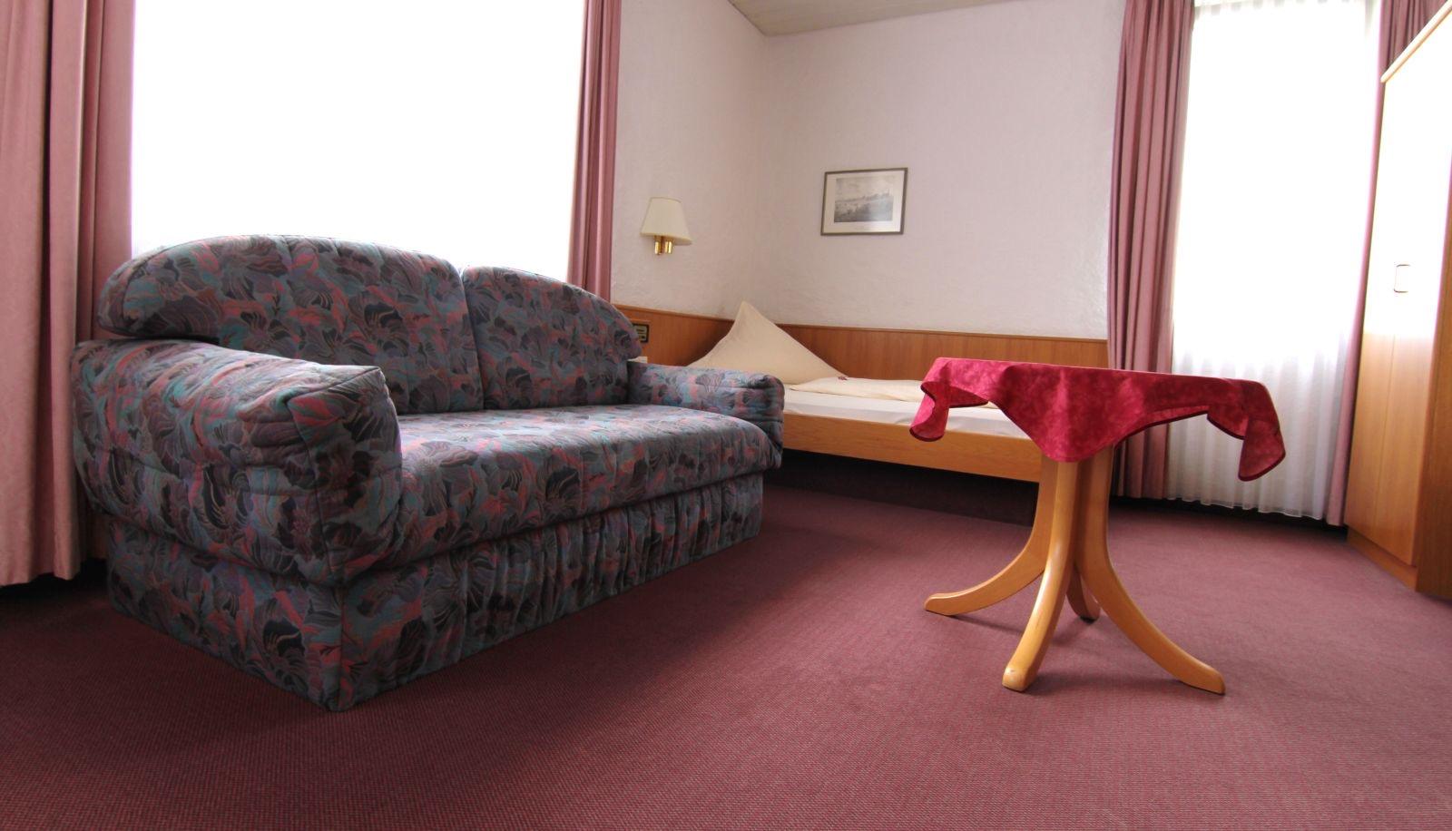 Dreibettzimmer mit Sofa, Einzelbett und rundem Tisch am Fenster